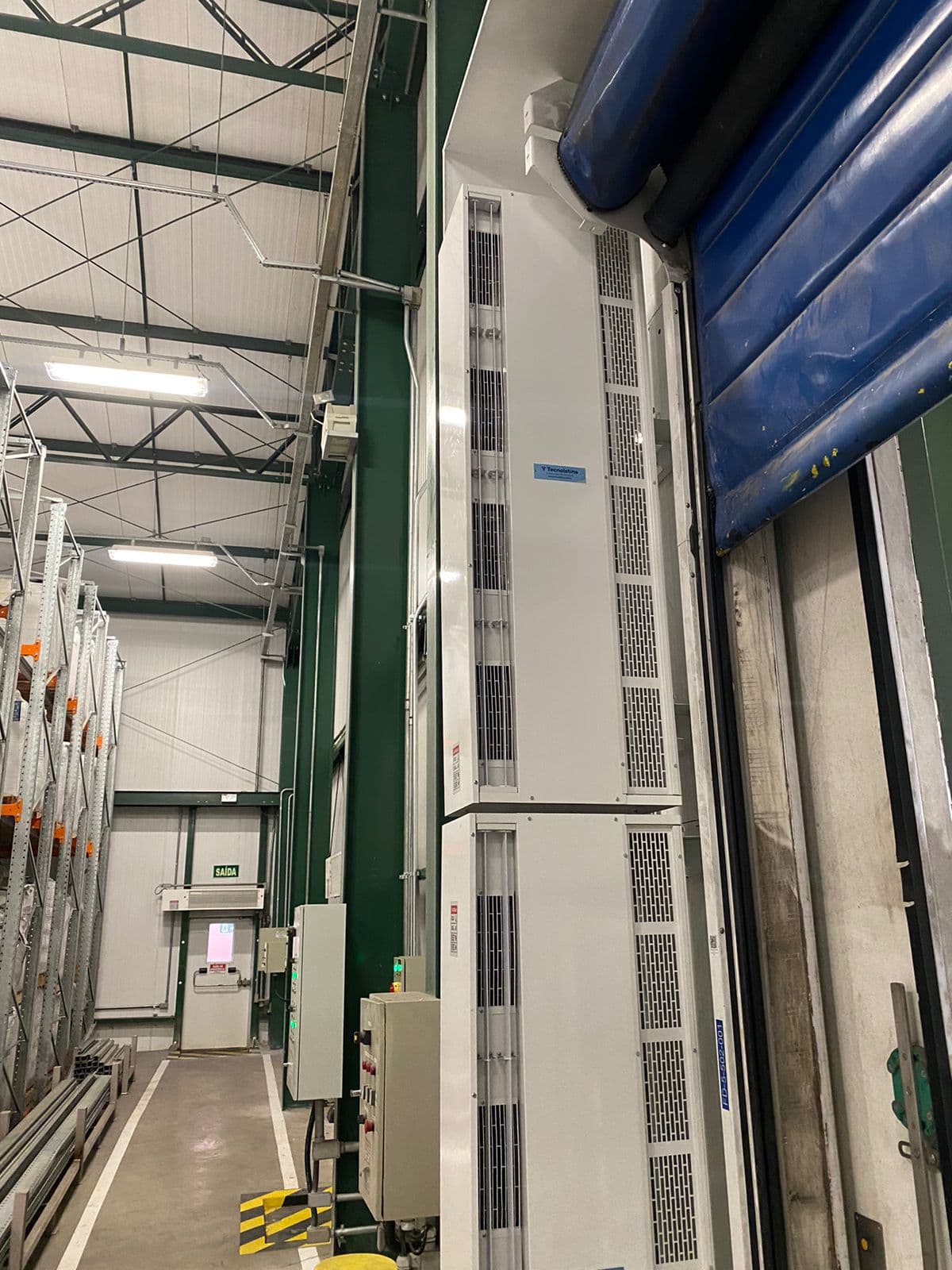 Módulos de cortina de ar vertical ao lado de porta rápida azul em galpão com estrutura verde